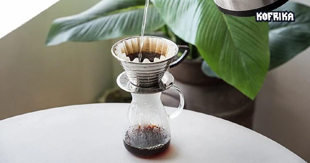  A glass jar of coffee with a handle. A filter is on top of the glass jar with filter paper inside. A silver pot is pouring water into the filter to brew the coffee.